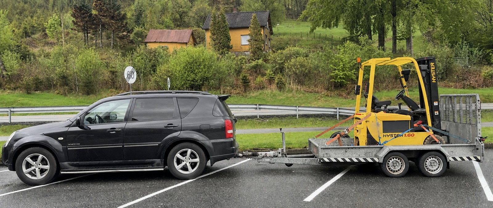 Svart SUV med maskinhenger og gul Hyster-truck under transport
