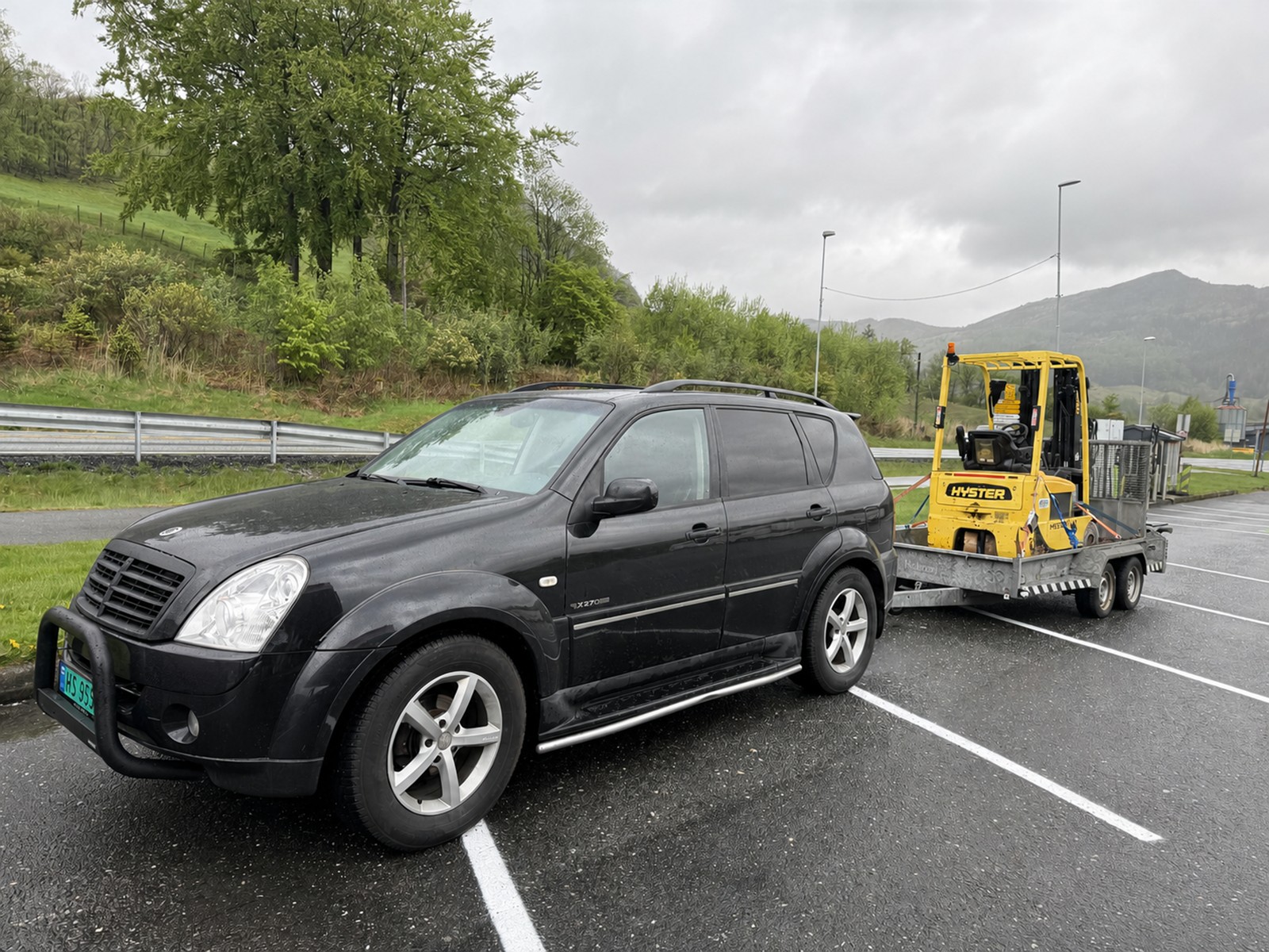 Svart SUV med gul Hyster-truck på maskinhenger i parkeringsområde